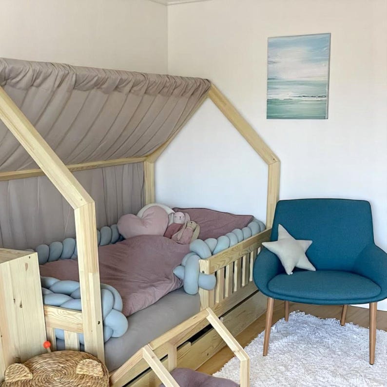 May include: A wooden framed children's bed with a canopy, featuring a pink duvet and a heart-shaped pillow. A blue armchair with a star cushion is next to the bed. A painting of the sea hangs on the wall. The room has a soft, inviting atmosphere.