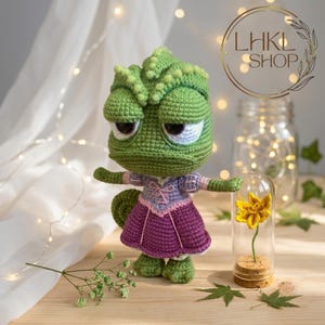 May include: A handmade green crochet chameleon doll wearing a purple and pink dress. The doll has a grumpy expression and is posed with its arms out. A yellow flower in a glass dome sits nearby. The shop logo "LHKL SHOP" is in the background.