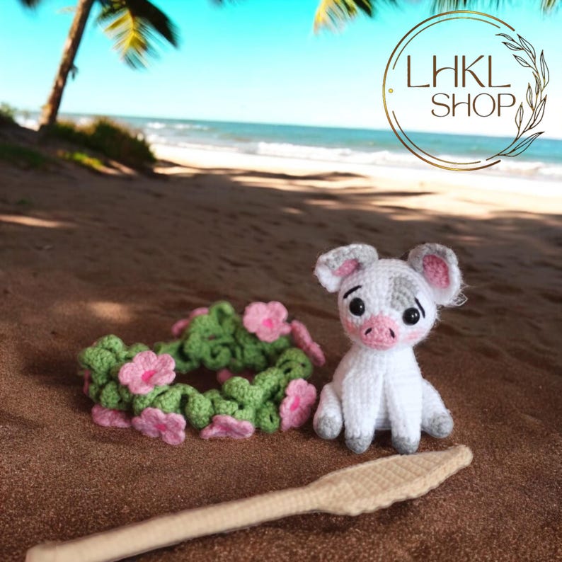 May include: A white crocheted pig with gray accents wearing a pink and green flower lei. The pig is sitting on a sandy beach with a wooden paddle in the foreground. The background shows a blue ocean and palm trees.