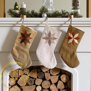 Può includere: Tre calze di Natale con motivi a stella sono appese a un caminetto bianco. Le calze sono nei toni del beige e del bianco, con disegni a stella marroni, beige e rossi. Il caminetto è decorato con verde e uno specchio.