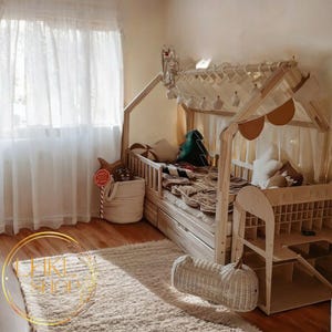 May include: A wooden bed frame shaped like a house with a white canopy and a white and brown patterned blanket. The bed has a built-in storage drawer and a ladder on the side. The bed is in a room with a white curtain and a rug on the floor.