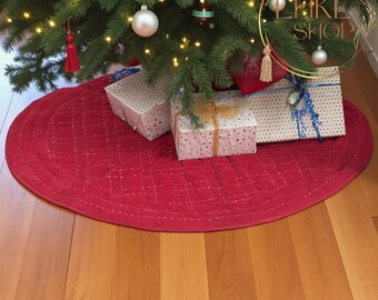 May include: A red quilted Christmas tree skirt with a stitched diamond pattern. The skirt is under a decorated Christmas tree with ornaments and wrapped gifts.