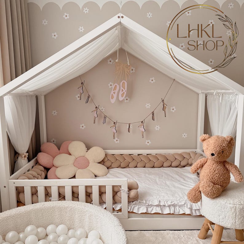 May include: A white canopy bed with a brown braided bumper and a white ruffled skirt. The bed has a pink flower-shaped pillow and a brown teddy bear sitting on a white stool. The wall behind the bed is decorated with pink flowers and a string of ballerina figurines.