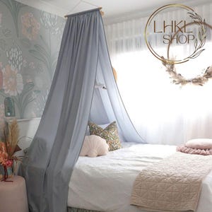 May include: A light gray canopy bed curtain with a white and floral patterned pillow. The bed has a white duvet and a pink quilt.