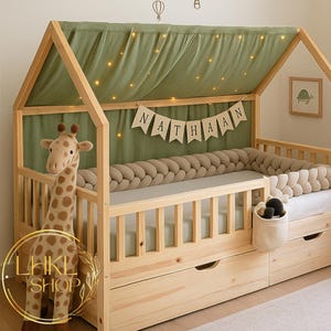 May include: A wooden house-shaped bed frame with a green canopy and fairy lights. The bed has a braided bumper, a giraffe plush toy, and a banner that says "NATHAAN". The bed also has two drawers.