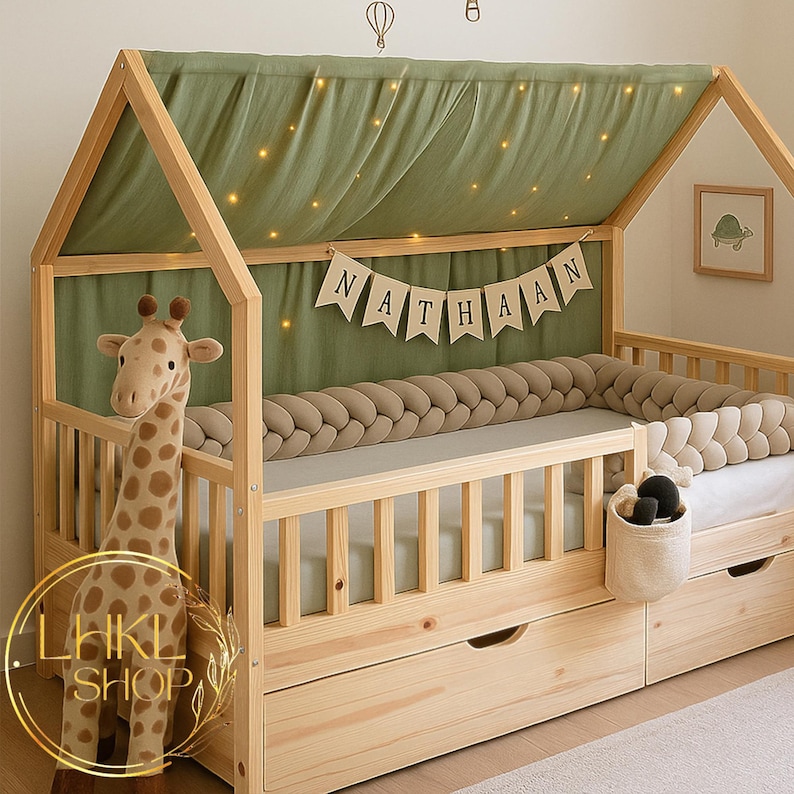 May include: A wooden house-shaped bed frame with a green canopy and fairy lights. The bed has a braided bumper, a giraffe plush toy, and a banner that says "NATHAAN". The bed also has two drawers.