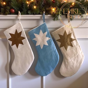 Peut inclure: Trois bas de Noël blancs, bleu clair et crème, chacun orné d'une étoile. Ils sont suspendus à une cheminée blanche décorée de verdure et de lumières, créant une scène festive pour les fêtes.