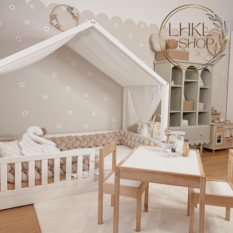 May include: A white canopy bed with a white and beige padded headboard and a white swan pillow. The bed is in a room with a light brown wall with white flower decals and a white scalloped border. There is a small wooden table and two chairs in the foreground. The text "LHK SHOP" is visible on a shelf in the background.