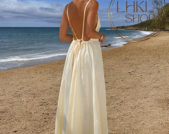 May include: A white maxi dress with a low back and spaghetti straps. The dress is made of a lightweight fabric and has a flowing silhouette. The dress is photographed on a beach.