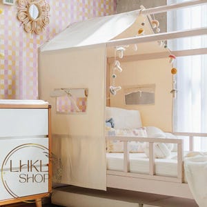 May include: A white canvas tent canopy for a bed, with a pink wooden frame and a white mattress. The tent has a window and a small flap that can be opened and closed. There are several felt mushroom decorations hanging from the frame.