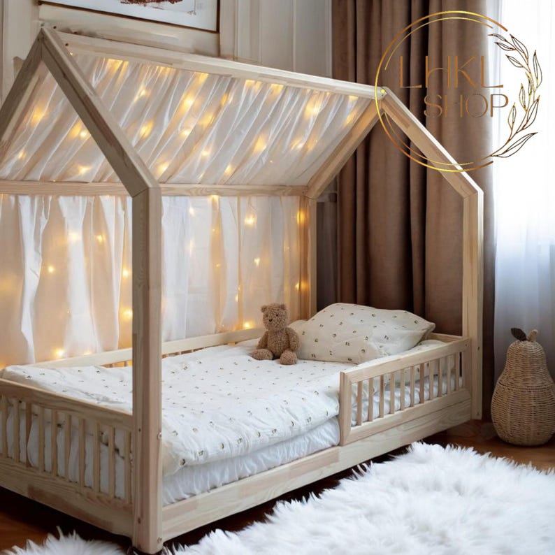 May include: A wooden toddler bed with a white canopy and string lights. The bed has a white mattress and a teddy bear on top. The bed is in a room with a white rug and brown curtains.