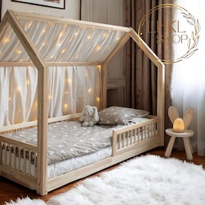 May include: A wooden house-shaped bed frame with a white canopy and a gray and white patterned mattress. The bed has a built-in guardrail and is decorated with fairy lights. A white furry rug is on the floor next to the bed.