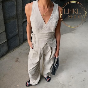 May include: A beige linen wrap jumpsuit with a V-neckline and a wide leg. The jumpsuit has a tie at the waist and a black handbag.