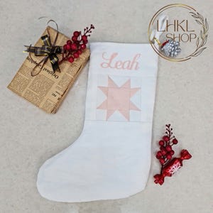 K&ouml;nnte beinhalten: Wei&szlig;er Weihnachtssocken mit einem rosafarbenen Stern-Patchworkmuster und dem Namen "Leah" auf der Vorderseite gestickt.