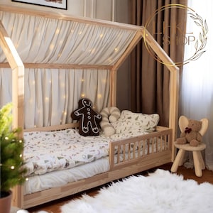 May include: A wooden house-shaped bed frame with a white canopy and a white and green patterned bedding. The bed has a safety rail and a gingerbread man pillow. A teddy bear sits on a wooden stool.