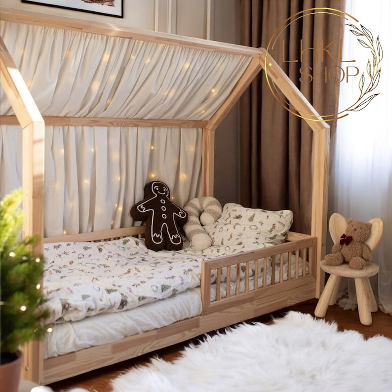 May include: A wooden house-shaped bed frame with a white canopy and a white and green patterned bedding. The bed has a safety rail and a gingerbread man pillow. A teddy bear sits on a wooden stool.