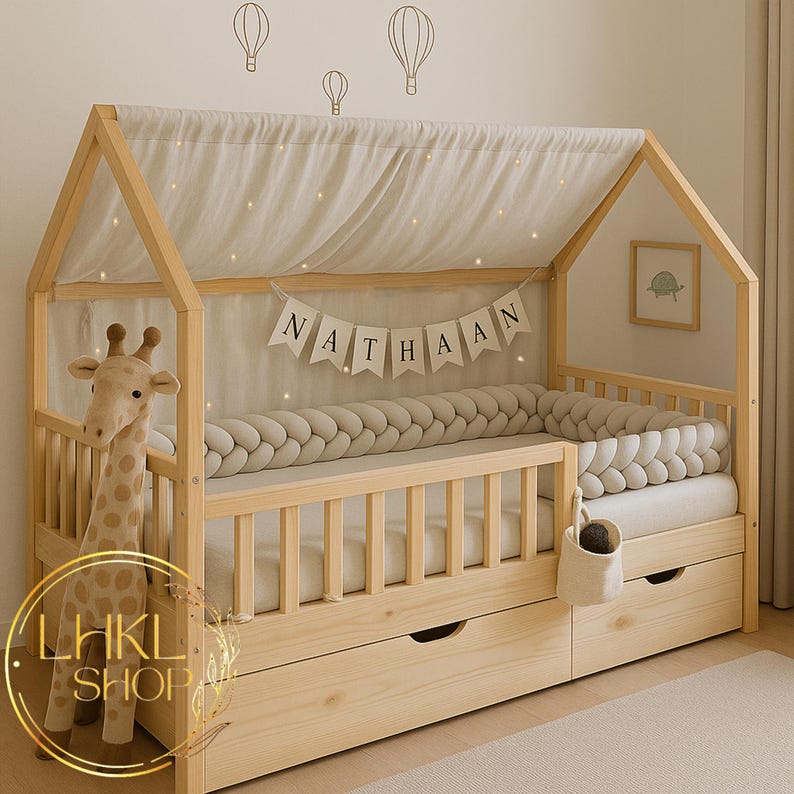 May include: A light wood toddler bed with a house-shaped canopy. The bed features a braided bumper, a banner reading "NATHAAN", and two drawers. A stuffed giraffe stands beside the bed.