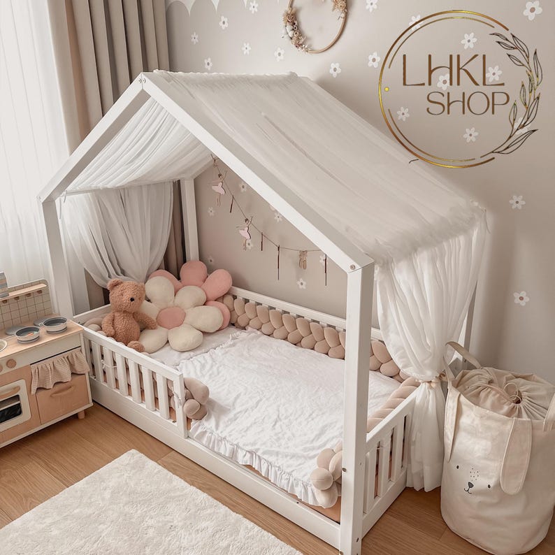 May include: White wooden canopy bed frame with a white canopy and a white and beige padded headboard. The bed is decorated with a pink flower pillow, a teddy bear, and a beige braided rope. The bed is in a room with a white wall and a white rug.