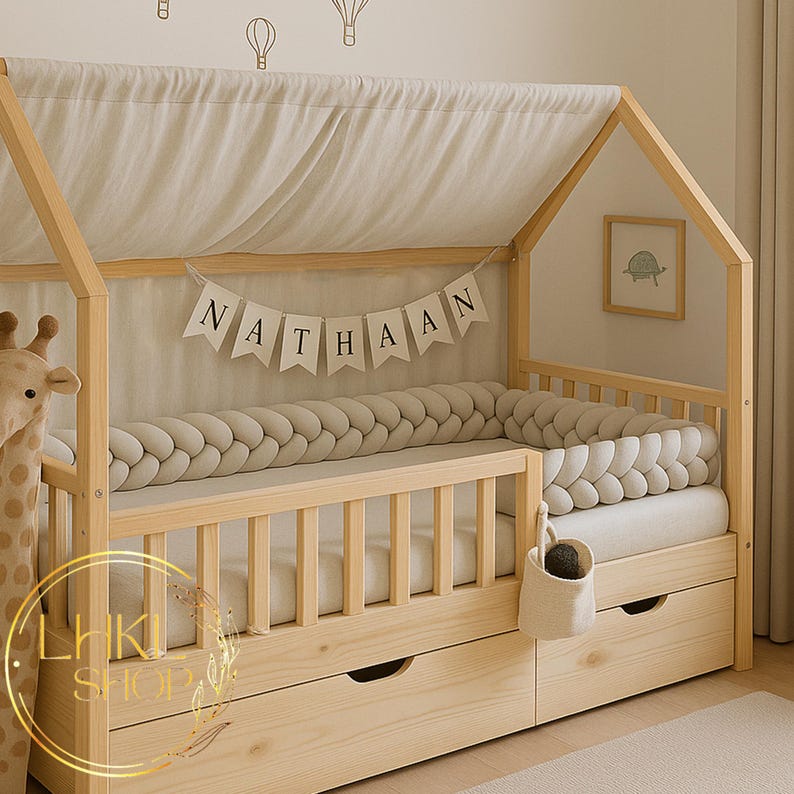 May include: A wooden bed with a house-shaped frame, featuring two drawers and a braided bumper. The bed has a canopy and a banner that reads "NATHAAN". A soft toy giraffe is on the left.