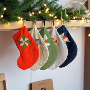 May include: Five Christmas stockings in various colors: red, cream, green, white, and navy blue. Each stocking features a geometric star design in complementary colors. They hang from a wooden shelf decorated with greenery and fairy lights.
