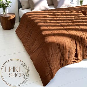 May include: A brown quilted bedspread on a bed with neutral-colored pillows. The bedspread has a textured appearance and is illuminated by sunlight. A wooden side table with a vase of flowers is visible in the background. The logo "LHKL SHOP" is in the lower left corner.