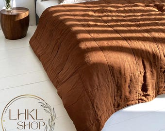 Può includere: Un copriletto trapuntato marrone su un letto con cuscini dai colori neutri. Il copriletto ha un aspetto strutturato ed è illuminato dalla luce solare. Un tavolino in legno con un vaso di fiori è visibile sullo sfondo. Il logo "LHKL SHOP" è in basso a sinistra.