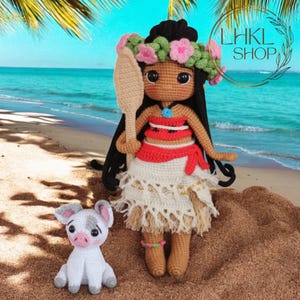 May include: A crocheted doll with brown hair wearing a flower crown and a red and white outfit, holding a wooden paddle. The doll is standing on a sandy beach with a small white crocheted pig beside it.