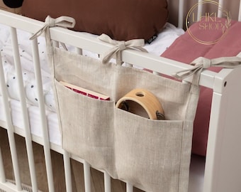 May include: A beige linen crib side organizer with two pockets. The organizer is hanging on a white crib rail. The pockets are filled with a book and a wooden toy.