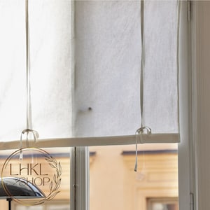 May include: White linen Roman shade with white ties, pulled up to reveal a window with a view of a building.