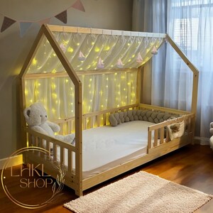 May include: A wooden house-shaped bed frame with a white canopy and string lights. The bed has a white mattress and a gray pillow. A teddy bear sits on the bed. The bed is in a room with a light brown rug and a white wall.