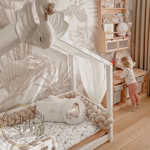 May include: A white canopy bed with a beige braided rope bumper and a white and gray floral bedding set. A stuffed white swan hangs from the canopy. A small child is standing next to a wooden shelf with books and baskets.