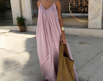 May include: A light purple maxi dress with spaghetti straps and a V-neck. The dress is flowing and loose-fitting. A large woven tote bag is held in the hand. The image is taken outdoors.