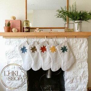 K&ouml;nnte beinhalten: F&uuml;nf wei&szlig;e Weihnachtsstr&uuml;mpfe mit bunten Sternmotiven h&auml;ngen an einem Holzmantel &uuml;ber einem Kamin. Die Str&uuml;mpfe haben einen Stern in den Farben Blau, Braun, Rot, Gelb und Gr&uuml;n.
