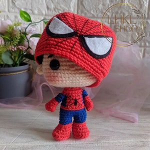 May include: A handmade crochet Spider-Man doll. The doll features a red and black Spider-Man mask with white eye accents, a red and blue suit, and red boots. The doll is standing on a white surface. The doll is a handmade item.