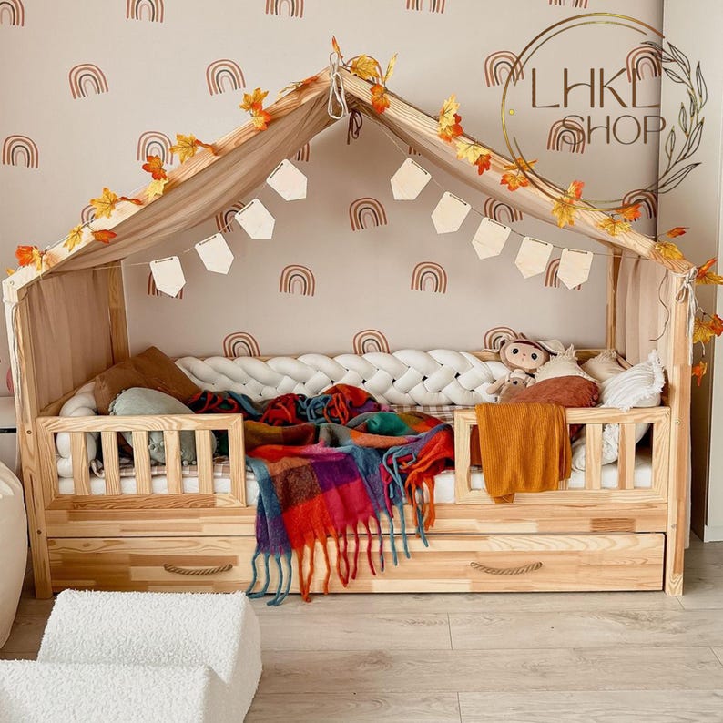 Pu&ograve; includere: Un lettino in legno per bambini piccoli con baldacchino e un materasso intrecciato bianco. Il letto ha un cassetto di stoccaggio ed &egrave; decorato con foglie autunnali e adesivi murali a forma di arcobaleno. Il testo "LHK SHOP" &egrave; visibile nell'immagine.