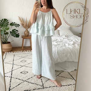 May include: A light blue linen pajama set. The set includes a camisole top with ruffle detailing and wide-leg pants with a drawstring waist. The outfit is displayed in a mirror, with a neutral-toned bedroom in the background.