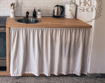 May include: A white kitchen cabinet with a wooden countertop and a white ruffled fabric skirt. The cabinet has a stainless steel sink and a black stovetop.