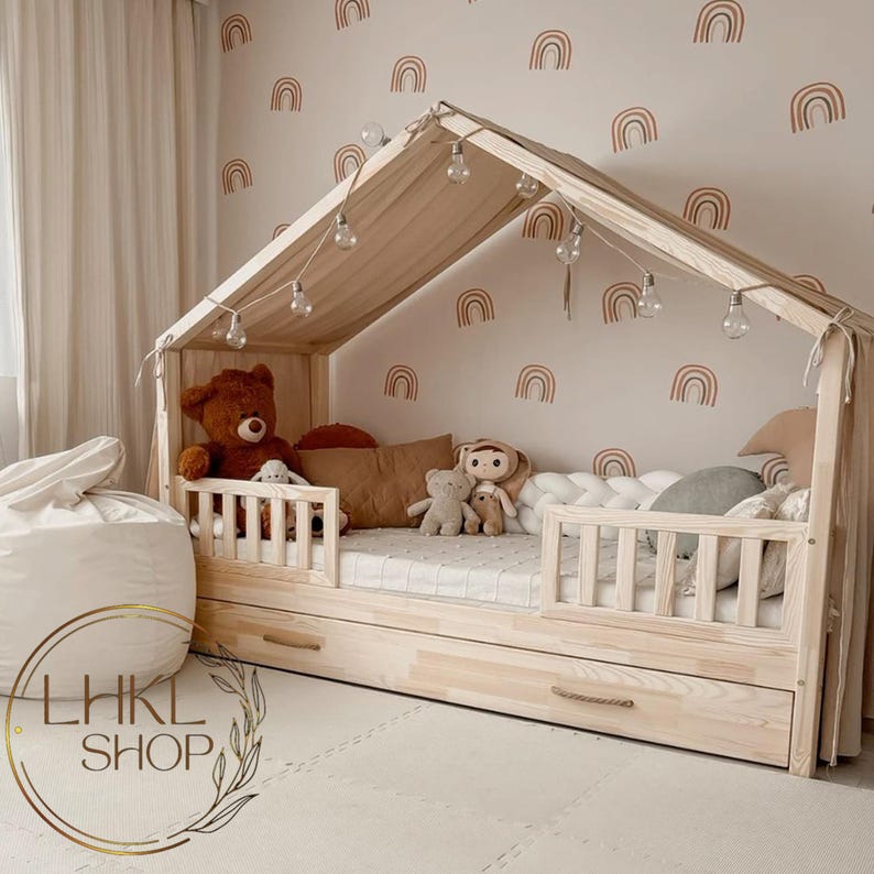 Pu&ograve; includere: Un letto in legno a forma di casa con un materasso bianco e un pouf bianco e soffice. Il letto ha un piumone bianco e grigio a fantasia e un cuscino bianco e soffice. La parete dietro il letto &egrave; decorata con adesivi a forma di arcobaleno.