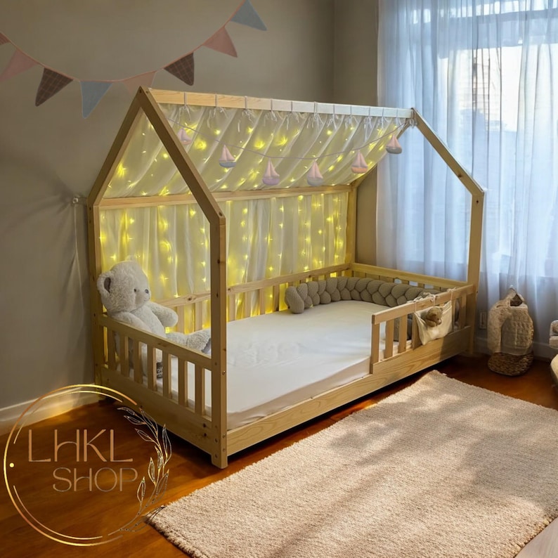 May include: A wooden bed frame shaped like a house with a white canopy and string lights. The bed has a white mattress and a gray pillow. The bed is in a room with a white rug and a window with white curtains.