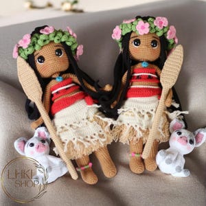 May include: Two crocheted dolls dressed as Moana from the Disney movie Moana. The dolls are wearing flower crowns, red and white tops, and brown skirts. They are holding wooden paddles. There is a small white crocheted pig next to the dolls.