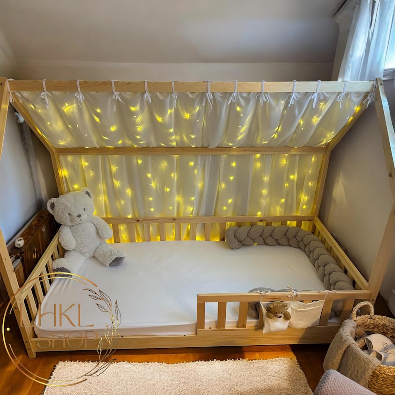 May include: A wooden bed frame with a white canopy and string lights. The bed has a white mattress, a gray braided pillow, and a white teddy bear. The bed is decorated with a white and green floral wreath.