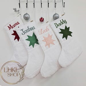 K&ouml;nnte beinhalten: Vier wei&szlig;e Weihnachtsstr&uuml;mpfe mit personalisierten Namen: Mama, Joshua, Leah und Daddy. Jeder Strumpf hat einen Stern-Patch in einer anderen Farbe: rot, gr&uuml;n, rosa und gr&uuml;n.