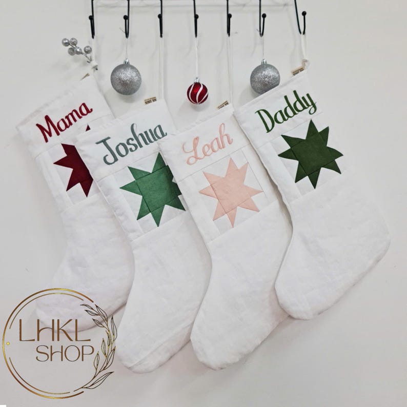 K&ouml;nnte beinhalten: Vier wei&szlig;e Weihnachtsstr&uuml;mpfe mit personalisierten Namen: Mama, Joshua, Leah und Daddy. Jeder Strumpf hat einen Stern-Patch in einer anderen Farbe: rot, gr&uuml;n, rosa und gr&uuml;n.