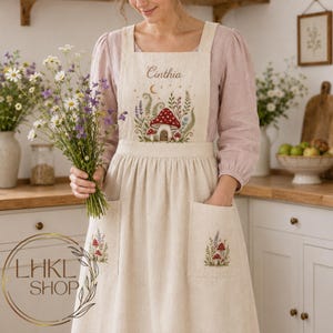 Op de afbeelding: Een beige linnen schort met een geborduurd ontwerp van een rood paddenstoelhuisje, wilde bloemen en een maansikkel. De naam Cinthia is bovenaan geborduurd. Het schort heeft twee voorzakken, elk versierd met kleine paddenstoelmotieven.