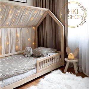 May include: A wooden house-shaped bed frame with a white canopy and string lights. The bed has a gray and white patterned bedding and a gray stuffed bunny. A wooden stool with a bunny-shaped nightlight sits next to the bed.