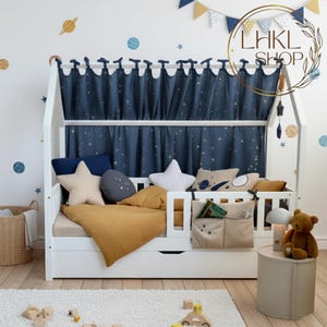 May include: A white wooden house bed with a navy blue star-patterned curtain. The bed is decorated with star and planet-shaped pillows, a mustard-colored blanket, and a small side table with a teddy bear.