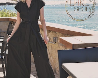 Puede incluir: Un mono negro de pierna ancha con cuello, mangas cortas y cinturón de lazo. El mono lo lleva puesto una persona de pie en un patio con vistas al mar.