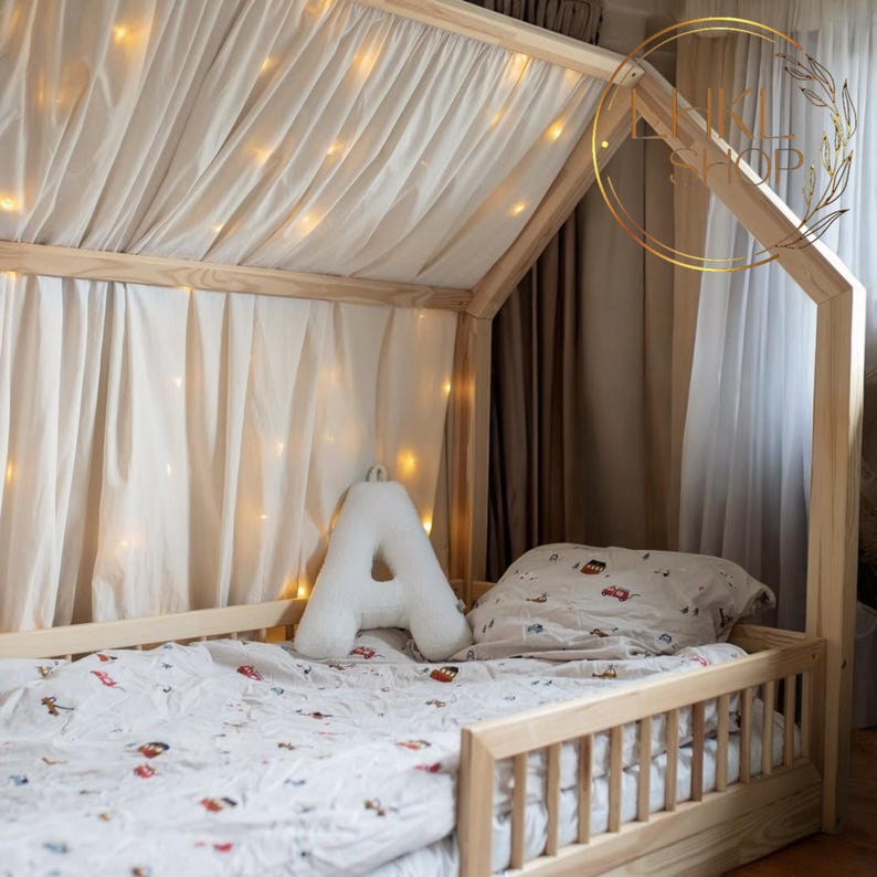 May include: A wooden house-shaped bed frame with a white canopy and string lights. The bed has a white pillow and a white letter A pillow. The bed has a white and gray patterned sheet with red and blue accents.