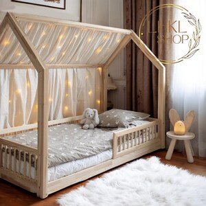May include: A wooden house-shaped bed frame with a white canopy and string lights. The bed has a gray and white patterned bedding and a small stuffed bunny on the bed. The bed is on a white fluffy rug.