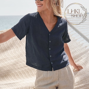 Puede incluir: Una camisa de lino azul marino con botones, cuello en V y mangas cortas. La camisa la lleva puesta una persona con el pelo rubio y se combina con pantalones a rayas.